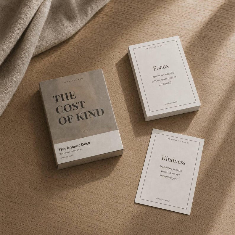 May include: A set of The Anchor Deck cards with a neutral color palette. The box reads "THE COST OF KIND" and the cards display the words "Focus" and "Kindness" with accompanying text. The cards are on a wooden surface.