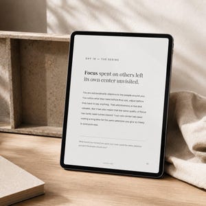 May include: A black tablet displaying text about focus and attention. The screen shows the title "DAY 16 - THE SEEING" and the phrase "Focus spent on others left its own center unvisited." The tablet is on a wooden surface.