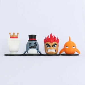 May include: Four cartoon character figurines. The first is a white cylinder with a gold crown. The second is a blue whale with a top hat and monocle. The third is a fiery-haired character with an angry face. The fourth is an orange fish.