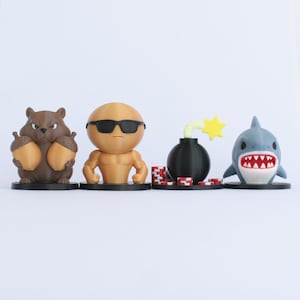May include: A collection of four whimsical figurines. A brown squirrel holding acorns, a muscular figure wearing sunglasses, a black bomb with a yellow star, and a blue shark with a red and white mouth.