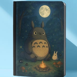 Puede incluir: Un cuaderno con una ilustración fantástica de una criatura grande, gris y peluda con vientre blanco, sentada cerca de una fogata bajo la luna llena. También hay una criatura blanca más pequeña y setas. El fondo representa una escena de bosque.