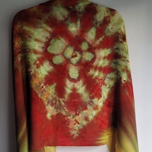 Könnte beinhalten: Ein Batik-Schal in Rot-, Gelb- und Grüntönen hängt vor einem weißen Hintergrund. Der Schal hat ein zentrales Design mit einem blütenartigen Muster. Die unteren Kanten haben Fransen.