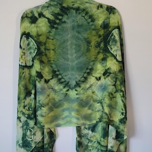 Könnte beinhalten: Ein grüner Schal mit Batikmuster und Fransen. Der Schal ist hauptsächlich in Grüntönen gehalten, mit helleren Gelbgrün- und dunkleren Waldgrüntönen. Der Stoff hat ein weiches, fließendes Aussehen, und die Fransen sind in einem dunkleren Grünton gehalten.