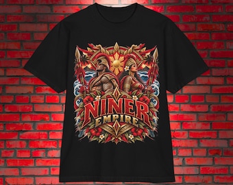 Puede incluir: Una camiseta negra de manga corta con un estampado gráfico de dos guerreros con cascos que sostienen lanzas, rodeados de follaje tropical, olas y un sol. El texto NINER EMPIRE está impreso en letras grandes de color rojo y dorado en el centro.