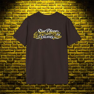 May include: A dark brown short-sleeve t-shirt featuring a graphic print on the chest. The design displays the text "San Diego" in a white cursive script above the word "CALIFAS" in a white block font, surrounded by yellow and white swirls.