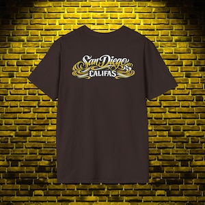 May include: A dark brown short-sleeve t-shirt featuring a graphic print on the chest. The design displays the text San Diego in a white cursive script font above the word CALIFAS in a white block letter font, accented with yellow swirls.