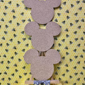 May include: Three cork coasters shaped like a mouse head are arranged on a small wooden easel. The background features a yellow pattern of bees and honeycomb. A small sign at the base of the easel reads "BeeCorked".