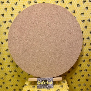 May include: A large, circular corkboard in natural brown. It's displayed on a small wooden easel, set against a yellow backdrop with a bee and honeycomb design. A business card with the text "BeeCorked" is visible.
