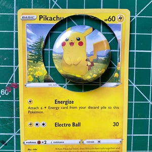 May include: A yellow Pikachu button badge on a Pikachu Pokémon trading card. The card features the text "Energize" and "Electro Ball". The badge shows a smiling Pikachu character.