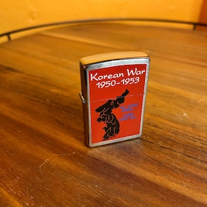 Puede incluir: Un encendedor plateado con un panel rojo que muestra un mapa negro de Corea. El texto "Korean War 1950-1953" está impreso encima del mapa. El encendedor está sobre una superficie de madera.