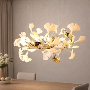 Puede incluir: Una lámpara de araña moderna con un marco dorado y detalles en forma de hojas. Las hojas son de color blanco crema, algunas con detalles dorados. La lámpara cuelga de finos cables, creando un efecto flotante. La lámpara de araña está suspendida sobre una mesa de madera.