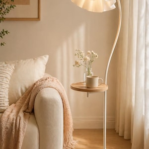 May include: A white floor lamp with a curved stem and a ruffled lampshade. The lamp has a small round wooden side table with a mug and a vase of flowers. The lamp is on a white marble base, next to a beige sofa with a pink throw.