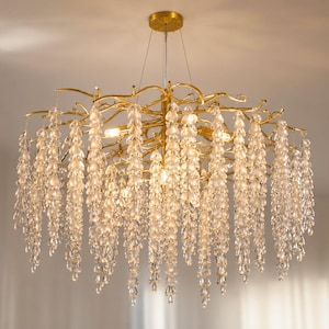 May include: A gold-toned chandelier featuring a branch-like frame with numerous cascading clear crystal teardrop-shaped pendants. The fixture is suspended by thin wires from a circular gold ceiling mount, creating a light-filled, organic display.