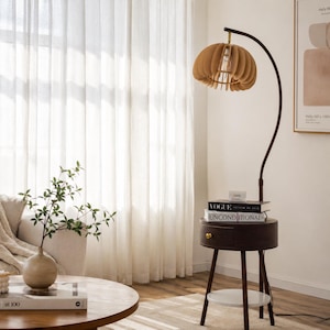 Könnte beinhalten: Eine braune Stehlampe mit einem einzigartigen, geschichteten Lampenschirm-Design. Die Lampe hat einen dunkelbraunen, gebogenen Metallarm und einen kleinen Beistelltisch mit einer Schublade. Bücher mit den Titeln "Vogue" und "Unconditional" sind auf dem Tisch gestapelt.