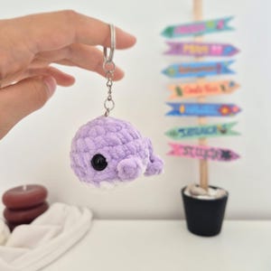 May include: A handmade, lavender crochet whale keychain with a black eye and white belly. The plush toy is attached to a silver keyring. The background includes a decorative signpost and candles.
