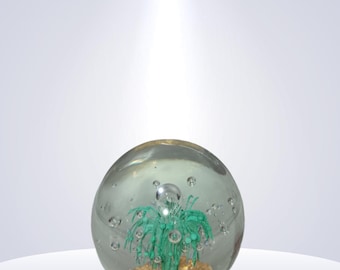 May include: A clear glass paperweight with a green plant design inside. The plant is set on a gold-colored base, and the glass sphere has several air bubbles. The paperweight is displayed on a white, curved surface.