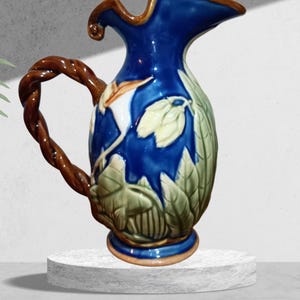 May include: A ceramic jug with a blue body and a brown, twisted handle. The jug features a floral design with green and white leaves. The rim and base have brown accents. The jug is displayed on a white plinth.
