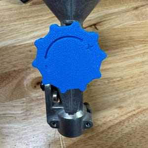 May include: A metal funnel with a blue, star-shaped adjustment knob. The knob has a textured surface and a curved line with a plus sign. The funnel is gray and has a conical shape.