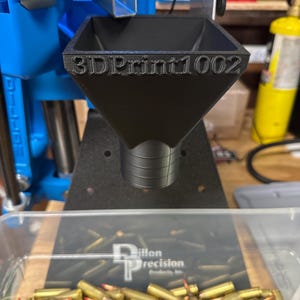 May include: Black 3D-printed funnel with the text "3DPrint1002" on the front. The funnel is positioned above a clear container filled with brass bullet casings. A blue reloading machine is in the background.