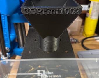 May include: Black 3D-printed funnel with the text "3DPrint1002" on the front. The funnel is positioned above a clear container filled with brass bullet casings. A blue reloading machine is in the background.