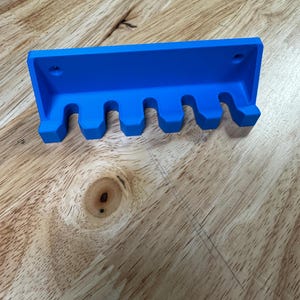 May include: A bright blue toothbrush holder with five curved slots. The holder is made of a solid material and has a rectangular back with two holes for mounting. The holder is designed to hold toothbrushes.