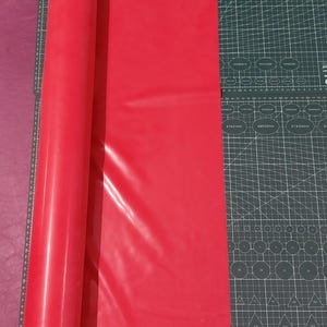Op de afbeelding: Een opgerolde baan helderrood materiaal naast een snijmat met rasterlijnen en een gele roterende snijder. De snijmat heeft afmetingen in centimeters.
