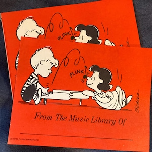 May include: Red cards featuring a Peanuts comic strip with Charlie Brown and Lucy playing a piano. The text "From The Music Library Of" is printed on the cards. The cards are on a blue surface.