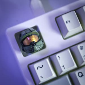 May include: A custom keycap in the shape of a green and gold helmet, possibly from a video game, fitted on a white keyboard. The keycap replaces a standard key, with the F1 key visible. The helmet features a gold visor.