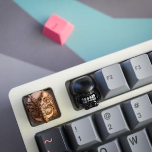 May include: Close-up of a white keyboard with custom keycaps. Two keycaps feature detailed alien-themed designs in brown and black. A pink keycap is visible in the background. The keyboard has gray keys with white lettering.