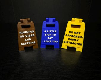 May include: Three colorful, miniature signboards with humorous text. One is brown and reads "RUNNING ON VIBES AND CAFFEINE." The second is blue and says "A LITTLE SIGN TO SAY I LOVE YOU." The third is yellow and says "DO NOT APPROACH: EASILY DISTRACTED."