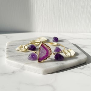 May include: A gold-toned bangle bracelet featuring a large, polished, semi-circular agate stone with pink and white bands. The bracelet is adorned with small purple amethyst stones and is displayed on a white marble surface.