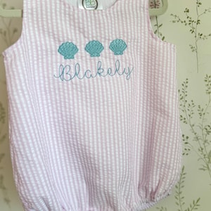 May include: Pink and white striped seersucker baby romper with three turquoise seashell appliques and the name "Blakely" embroidered in cursive. The romper has button closures on the shoulders and at the bottom.