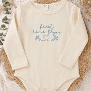 May include: A cream-colored long-sleeve bodysuit featuring blue embroidery on the chest that reads "first time flyer" with two small airplane icons connected by a heart-shaped flight path. The garment includes a snap closure at the neckline.