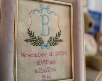 May include: A framed embroidered artwork with a pink and gold frame. The artwork features a blue letter "B" within a wreath, with the date November 11, 2024, time, weight (6lbs 7oz), and length (19 in) embroidered in pink.