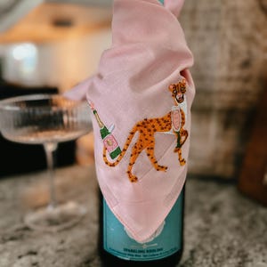 May include: A pink champagne bottle scarf with an embroidered cheetah design holding a champagne glass and bottle. The cheetah has orange spots and is wearing a white collar. The scarf is wrapped around a sparkling riesling bottle.