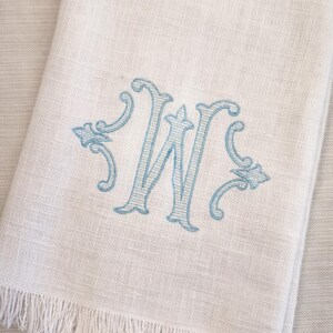 May include: White linen hand towel with a light blue embroidered letter "W". The towel has a fringed edge and a textured weave. The embroidered letter is in a decorative script style.