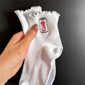 May include: White ribbed socks with a ruffled top edge. Each sock features an embroidered Coke can design in red and black. The socks are held against a dark background.