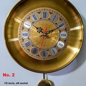 May include: A gold-coloured, all-metal wall clock with a pendulum. The clock face features Roman numerals and decorative accents. The clock is approximately 25 cm in diameter.