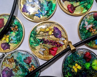 Puede incluir: Colgantes redondos de resina con bordes dorados, con flores secas y mariposas. Los colgantes están llenos de purpurina y colocados sobre un cordón negro. Las flores son rojas, amarillas y moradas.