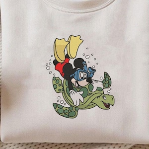 May include: Cream-colored sweatshirt featuring a cartoon Mickey Mouse character scuba diving on a green turtle. Mickey wears a red shirt, black shorts, and yellow flippers. The design includes bubbles and is set against a neutral background.