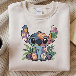 May include: Cream-colored sweatshirt featuring a colorful Stitch graphic with floral patterns. The design includes Stitch's large ears, eyes, and body, surrounded by tropical leaves. A coffee cup and notebook are in the background.