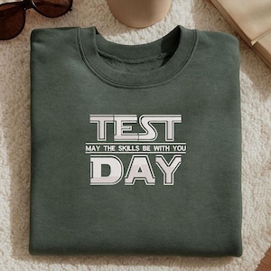 Puede incluir: Sudadera verde oscuro con las palabras "TEST DAY" en letras blancas. La frase "MAY THE SKILLS BE WITH YOU" está impresa debajo. La sudadera está doblada sobre una superficie blanca.