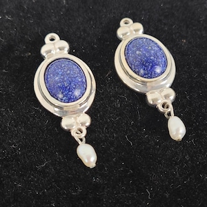 May include: Two silver-toned earrings with oval, deep blue lapis lazuli stones. Each earring has a small pearl drop. The earrings have a decorative top and bottom design. The background is a dark, textured surface.