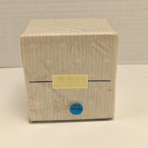 Puede incluir: Caja de perfume beige con un patrón repetido y la palabra "devotee" en un rectángulo amarillo. Una pegatina azul con el texto "5115 devotee" está en la parte inferior. La caja está envuelta en plástico transparente.