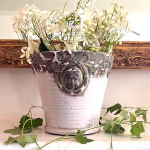 May include: A white ceramic planter with a decorative face and floral accents, filled with white flowers. The planter has a textured surface and is set against a gold-framed mirror.