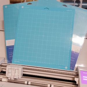 May include: Several light blue cutting mats with a grid pattern and the text "Light Grip" are stacked on top of a white cutting machine. The mats are designed for crafting projects and have a 12-inch cutting area.
