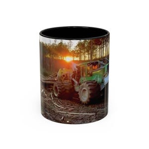 May include: A black and white ceramic mug featuring a vibrant image of a green logging machine in a forest at sunset. The mug's interior is black, and the exterior showcases the machinery and the natural environment.