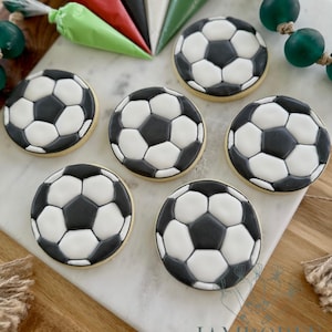 May include: Six soccer ball-shaped cookies with black and white icing, arranged on a marble surface. Piping bags filled with red, black, white, and green icing are visible in the background, along with green bead garland. The cookies have a yellow base.