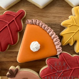 May include: A collection of decorated sugar cookies with a Thanksgiving theme. The cookies include a slice of pumpkin pie, red and yellow fall leaves, and a turkey. The cookies are arranged on a wooden surface.