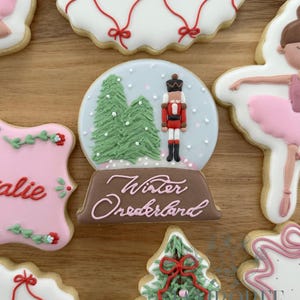 Op de afbeelding: Assortiment van versierde suikerkoekjes op een houten oppervlak. Koekjes zijn onder meer een sneeuwbol met een notenkraker, een kerstboom en een ballerina. Het sneeuwbolkoekje heeft de tekst "Winter Wonderland".